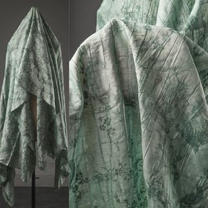 May include: A light green fabric drape featuring a dark green botanical print with branches, leaves, and flowers. The material has a crinkled, textured surface and is displayed on a stand, showing folds and drapes throughout the textile.
