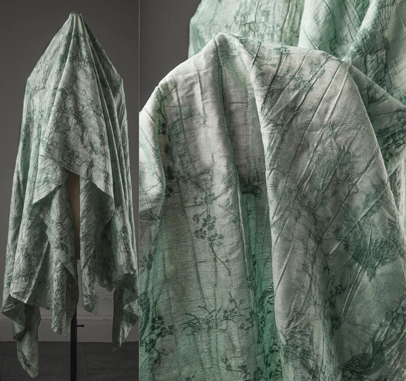 May include: A light green fabric drape featuring a dark green botanical print with branches, leaves, and flowers. The material has a crinkled, textured surface and is displayed on a stand, showing folds and drapes throughout the textile.