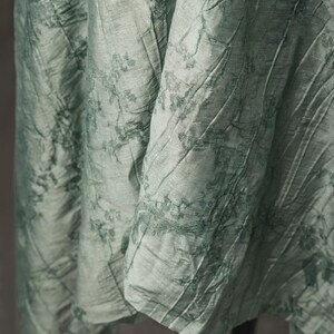 May include: A light sage green fabric with a crinkled texture and a darker green botanical print featuring delicate floral and leaf patterns. The material drapes in vertical folds, showcasing a subtle sheen and a complex, layered visual depth.