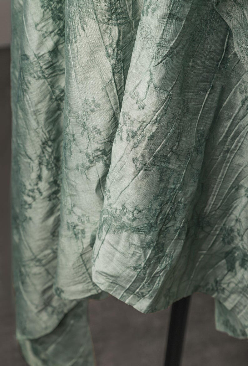 May include: A light sage green fabric with a crinkled texture and a darker green botanical print featuring delicate floral and leaf patterns. The material drapes in vertical folds, showcasing a subtle sheen and a complex, layered visual depth.