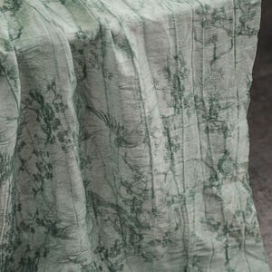 May include: A light green fabric table runner featuring a dark green printed pattern of trees, branches, and birds. The textile has a crinkled, textured surface with vertical folds and a semi-sheer appearance, draped to show its length and drape.