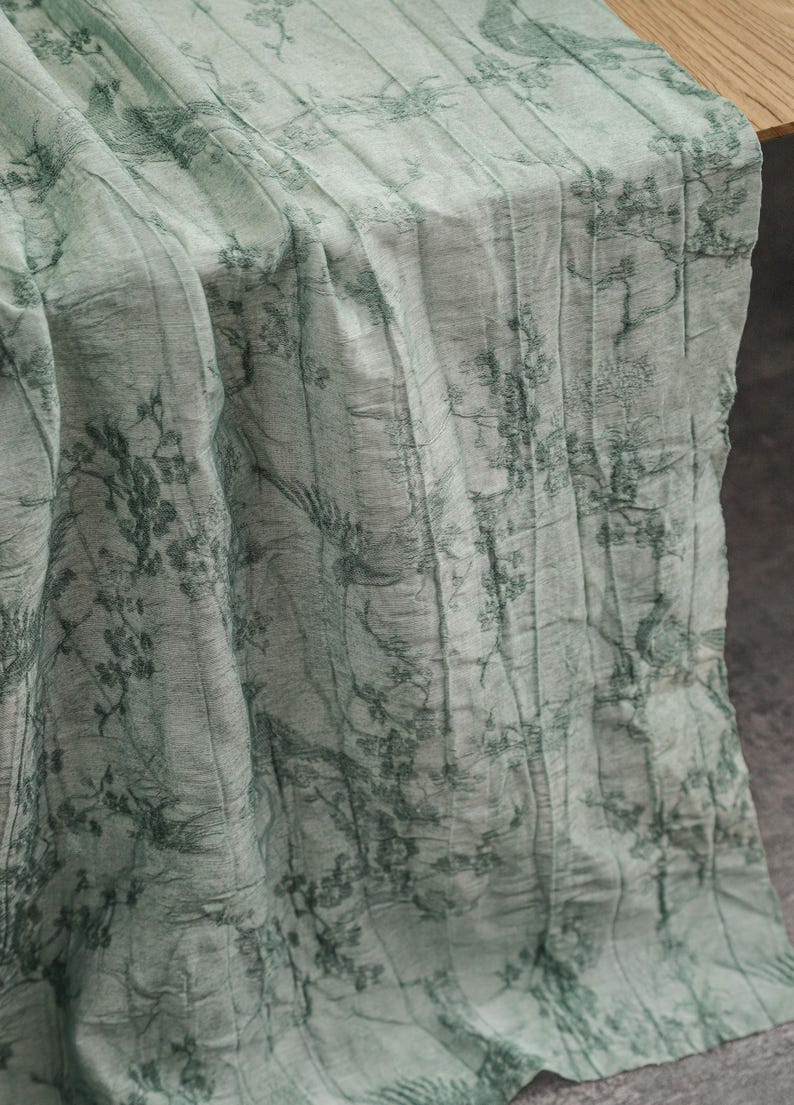 May include: A light green fabric table runner featuring a dark green printed pattern of trees, branches, and birds. The textile has a crinkled, textured surface with vertical folds and a semi-sheer appearance, draped to show its length and drape.