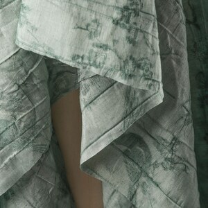 May include: A light sage green fabric featuring a dark green botanical print with intricate floral and vine patterns. The material has a visible textured weave and is draped in soft folds, creating a layered appearance with varying light and shadow.