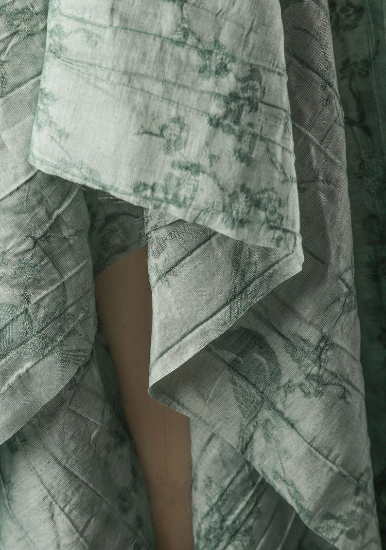 May include: A light sage green fabric featuring a dark green botanical print with intricate floral and vine patterns. The material has a visible textured weave and is draped in soft folds, creating a layered appearance with varying light and shadow.