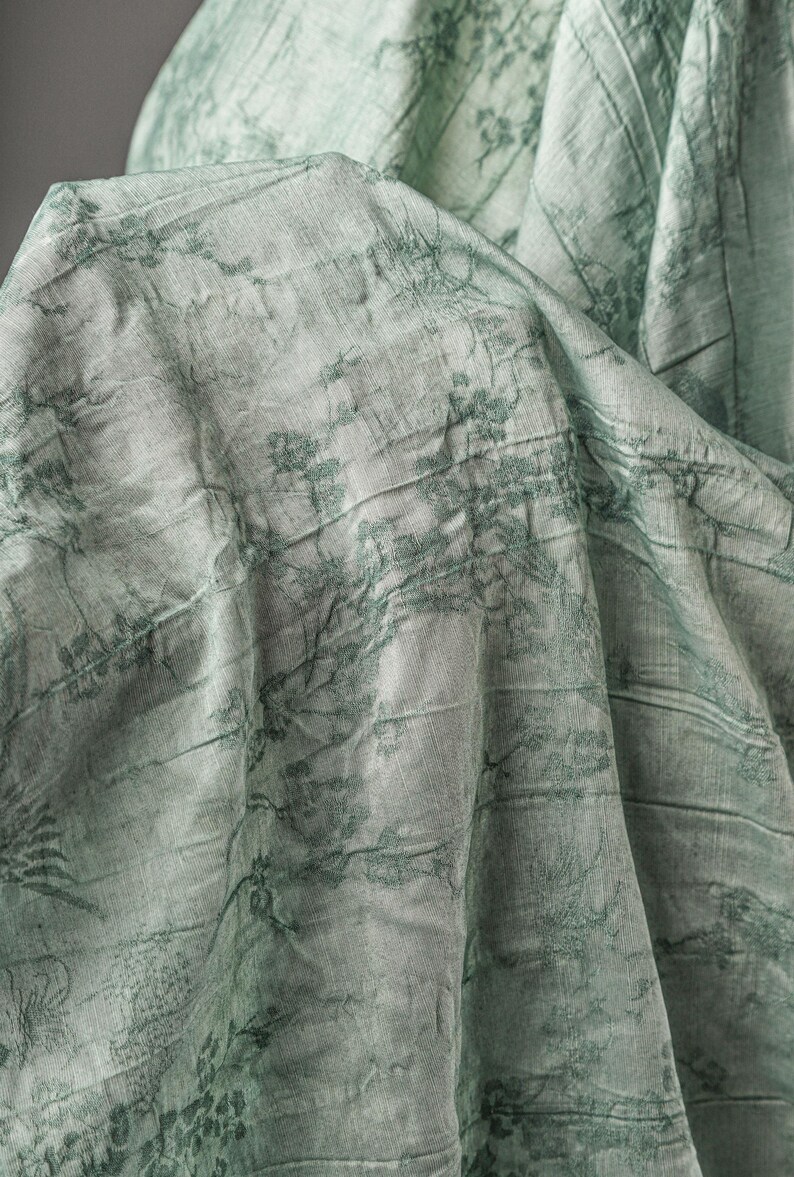 May include: A light mint green fabric featuring a dark green printed pattern of delicate branches and foliage. The material has a visible crinkled texture with folds and drapes that create depth and shadows across the surface of the textile.