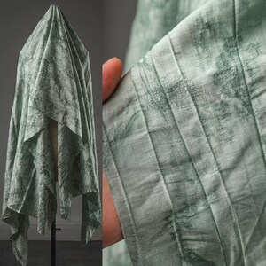 May include: A light sage green fabric featuring a textured, crinkled surface with a darker green abstract pattern. The material is draped in vertical folds, showcasing a semi-sheer quality and a fine, linear weave throughout the textile design.