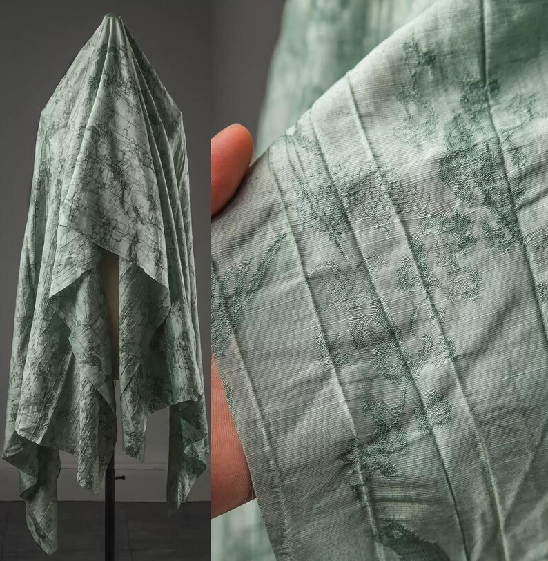 May include: A light sage green fabric featuring a textured, crinkled surface with a darker green abstract pattern. The material is draped in vertical folds, showcasing a semi-sheer quality and a fine, linear weave throughout the textile design.