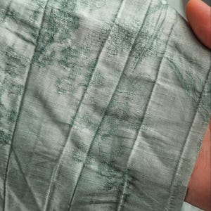 May include: A light sage green fabric featuring a subtle, darker green floral pattern. The material has a distinct pleated texture with vertical lines running throughout, creating a layered and dimensional appearance across the surface of the textile.