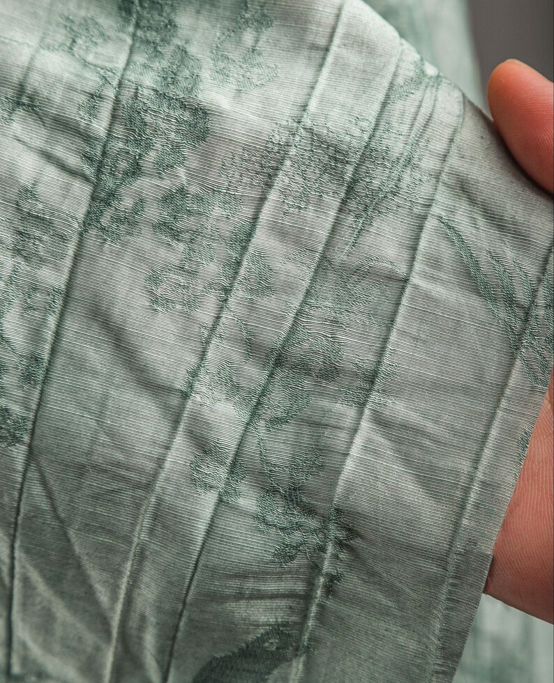 May include: A light sage green fabric featuring a subtle, darker green floral pattern. The material has a distinct pleated texture with vertical lines running throughout, creating a layered and dimensional appearance across the surface of the textile.
