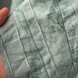 May include: A light sage green fabric featuring a series of vertical pleats and a darker green floral pattern. The material has a visible textured weave and a subtle sheen, with the pleats creating distinct lines across the surface of the textile.