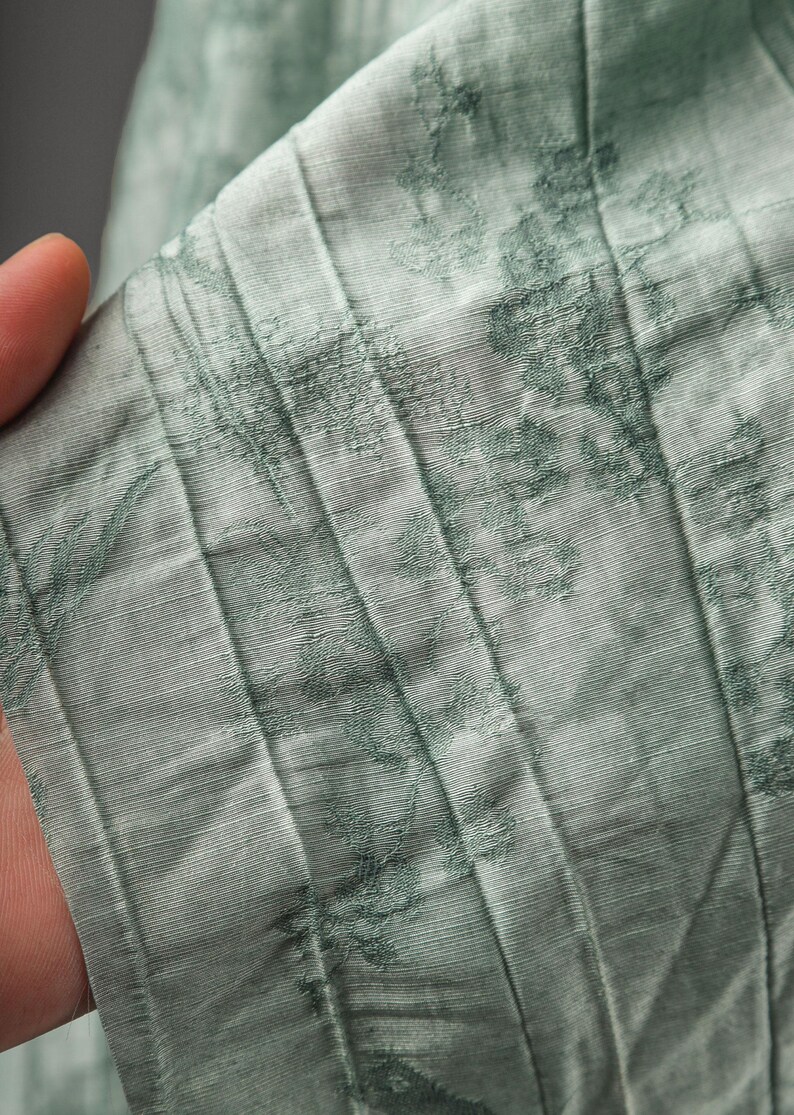 May include: A light sage green fabric featuring a series of vertical pleats and a darker green floral pattern. The material has a visible textured weave and a subtle sheen, with the pleats creating distinct lines across the surface of the textile.