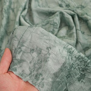 May include: A light sage green fabric featuring a dark green botanical print of branches and leaves. The material has a crinkled, textured surface and a semi-sheer quality, showing folds and drapes throughout the length of the textile.