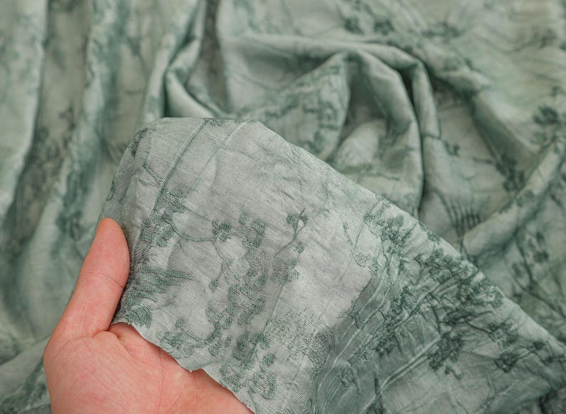 May include: A light sage green fabric featuring a dark green botanical print of branches and leaves. The material has a crinkled, textured surface and a semi-sheer quality, showing folds and drapes throughout the length of the textile.