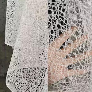 May include: Close-up of white lace fabric with a delicate, openwork design. The fabric has a net-like appearance with numerous small, circular openings. The texture is soft and intricate, suggesting a fine weave. The lace is likely used for clothing or decorative purposes.