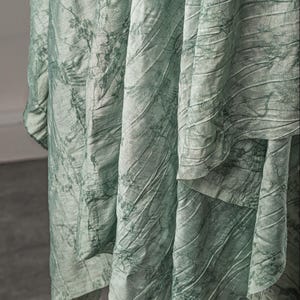 May include: A light sage green fabric with a crinkled, textured surface featuring a dark green botanical print of delicate branches and leaves. The material hangs in deep, vertical folds, showcasing the intricate pattern and the uneven, rumpled finish.
