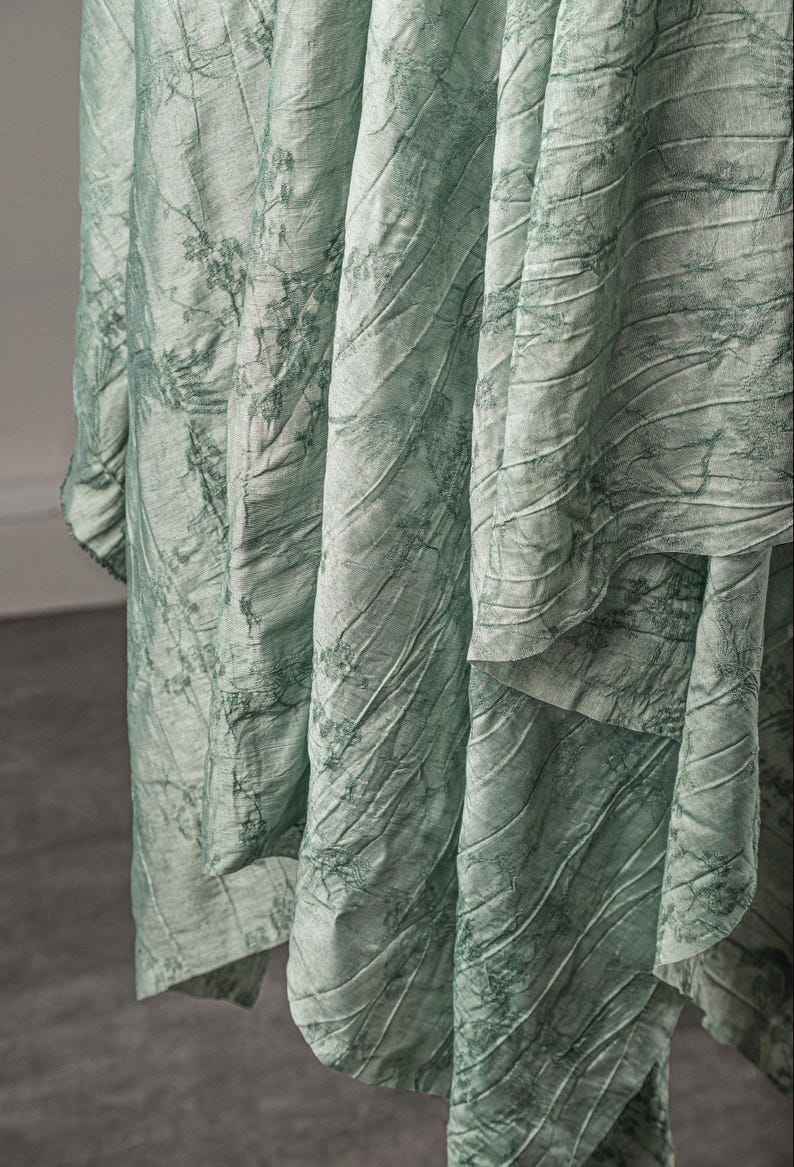 May include: A light sage green fabric with a crinkled, textured surface featuring a dark green botanical print of delicate branches and leaves. The material hangs in deep, vertical folds, showcasing the intricate pattern and the uneven, rumpled finish.