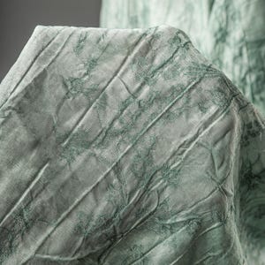 May include: A light sage green fabric featuring a crinkled, textured surface with a darker green botanical print. The material displays vertical folds and creases, creating a dimensional appearance across the draped, semi-sheer textile surface.
