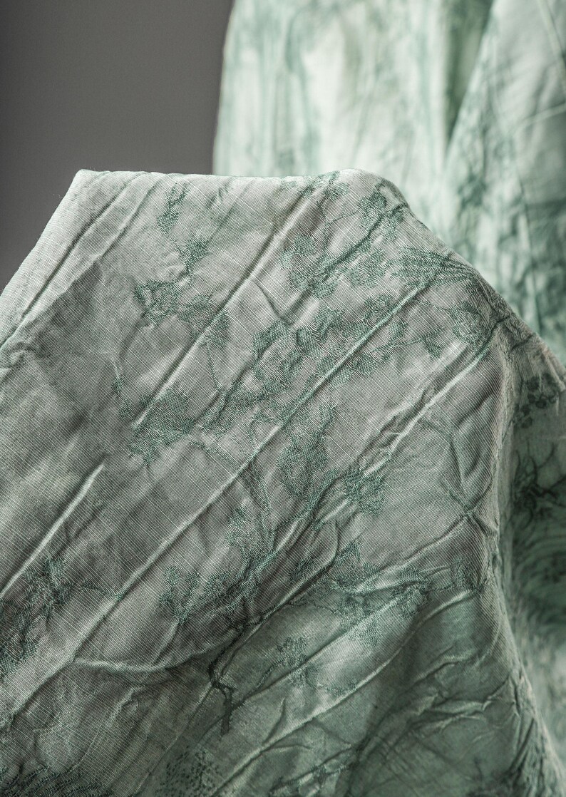 May include: A light sage green fabric featuring a crinkled, textured surface with a darker green botanical print. The material displays vertical folds and creases, creating a dimensional appearance across the draped, semi-sheer textile surface.