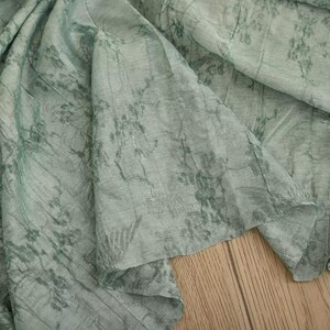 May include: A light sage green fabric featuring a dark green botanical print with intricate floral and vine patterns. The material has a visible woven texture with soft folds and drapes, creating a layered appearance across the surface.