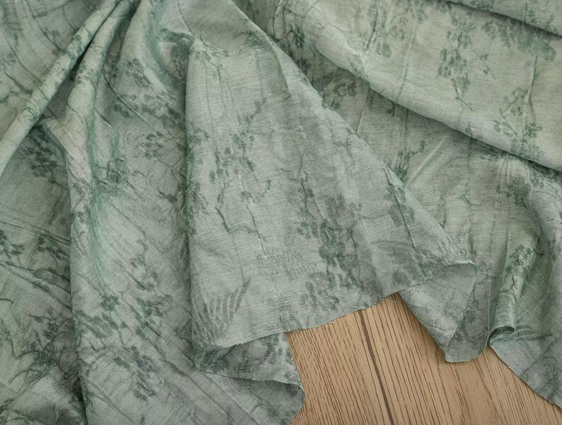 May include: A light sage green fabric featuring a dark green botanical print with intricate floral and vine patterns. The material has a visible woven texture with soft folds and drapes, creating a layered appearance across the surface.