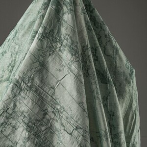 May include: A light sage green fabric draped in a conical shape, featuring a dark green printed pattern of intricate tree branches and foliage. The textile has a visible woven texture with subtle horizontal lines and soft, flowing folds.
