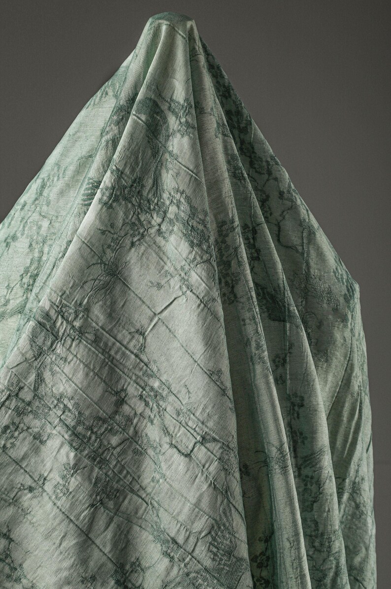 May include: A light sage green fabric draped in a conical shape, featuring a dark green printed pattern of intricate tree branches and foliage. The textile has a visible woven texture with subtle horizontal lines and soft, flowing folds.
