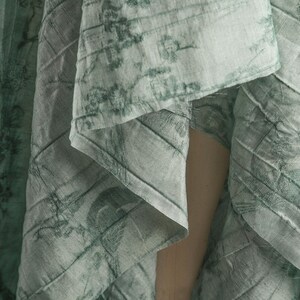 May include: A light green fabric with a darker green botanical print featuring birds and branches. The material has a visible quilted texture with diagonal stitching lines running across the surface, creating a layered and draped appearance.