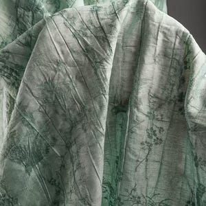 May include: A light green fabric featuring a dark green botanical print with detailed floral and branch motifs. The material has a distinct crinkled, textured surface with visible folds and creases that create depth across the draped textile.