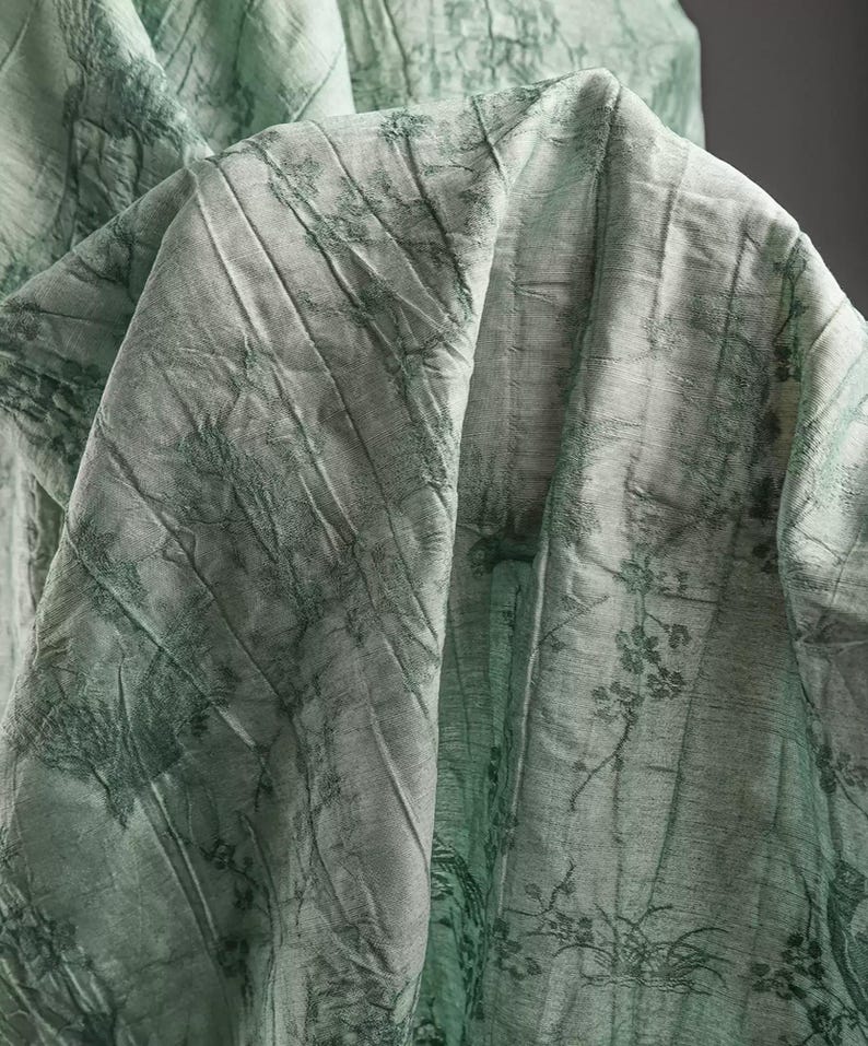 May include: A light green fabric featuring a dark green botanical print with detailed floral and branch motifs. The material has a distinct crinkled, textured surface with visible folds and creases that create depth across the draped textile.