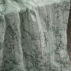 May include: A light sage green fabric runner featuring a dark green toile-style print of trees, branches, and birds. The material has a distinct vertical crinkled texture throughout, creating a series of parallel folds across the surface.