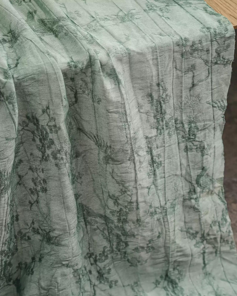 May include: A light sage green fabric runner featuring a dark green toile-style print of trees, branches, and birds. The material has a distinct vertical crinkled texture throughout, creating a series of parallel folds across the surface.