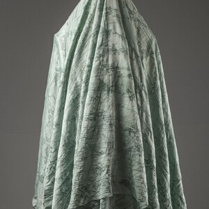 May include: A light mint green fabric draped vertically, featuring a dark green printed pattern of birds in flight and intricate botanical elements. The textile has a subtle crinkled texture throughout, creating deep folds and shadows.