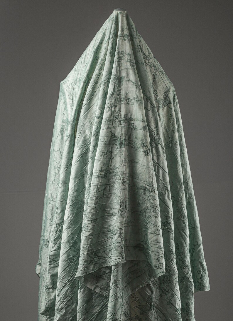 May include: A light mint green fabric draped vertically, featuring a dark green printed pattern of birds in flight and intricate botanical elements. The textile has a subtle crinkled texture throughout, creating deep folds and shadows.