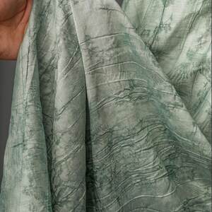 May include: A light sage green fabric featuring a textured, crinkled surface with a darker green botanical print of tree branches and foliage. The material is draped in soft folds, showcasing the intricate pattern and the subtle sheen of the textile.