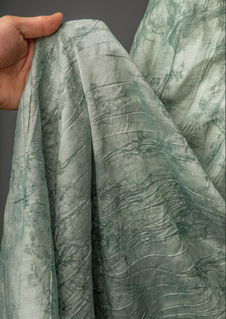 May include: A light sage green fabric featuring a textured, crinkled surface with a darker green botanical print of tree branches and foliage. The material is draped in soft folds, showcasing the intricate pattern and the subtle sheen of the textile.
