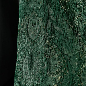 May include: Close-up of a textured green fabric with a paisley pattern. The fabric has a shimmering quality with gold accents, creating a luxurious and detailed appearance. The design features intricate floral and curved shapes.