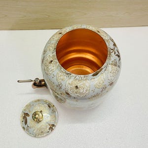 K&ouml;nnte beinhalten: Ein wei&szlig;-goldenes Kupferkessel mit floralem Muster und passendem Deckel. Das Innere des Kessels ist kupferfarben. Der Kessel hat einen Ausguss und einen Griff. Der Deckel ist daneben.