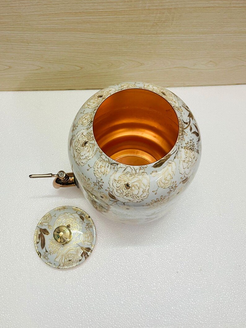 K&ouml;nnte beinhalten: Ein wei&szlig;-goldenes Kupferkessel mit floralem Muster und passendem Deckel. Das Innere des Kessels ist kupferfarben. Der Kessel hat einen Ausguss und einen Griff. Der Deckel ist daneben.
