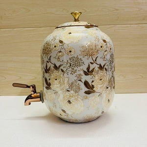 K&ouml;nnte beinhalten: Ein wei&szlig;er Getr&auml;nkespender aus Keramik mit einem floralen Muster in Gold und Braun. Er hat einen goldenen Deckel und einen kupferfarbenen Wasserhahn. Der Spender dient zum Ausschenken von Getr&auml;nken.