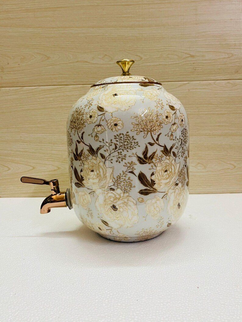 K&ouml;nnte beinhalten: Ein wei&szlig;er Getr&auml;nkespender aus Keramik mit einem floralen Muster in Gold und Braun. Er hat einen goldenen Deckel und einen kupferfarbenen Wasserhahn. Der Spender dient zum Ausschenken von Getr&auml;nken.