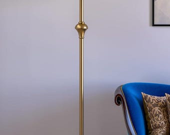 Könnte beinhalten: Eine Stehlampe mit einem hohen, schlanken, goldfarbenen Metallständer, der in der Mitte ein dekoratives, bauchiges Element aufweist. Die Lampe hat einen runden Fuß und einen breiten, konisch geformten Lampenschirm aus cremefarbenem Stoff.