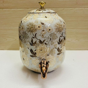 K&ouml;nnte beinhalten: Ein wei&szlig;er Getr&auml;nkespender aus Keramik mit einem floralen Muster in Gold und Braun. Er hat einen goldenen Deckel und einen kupferfarbenen Zapfhahn. Geeignet zum Servieren von Getr&auml;nken.