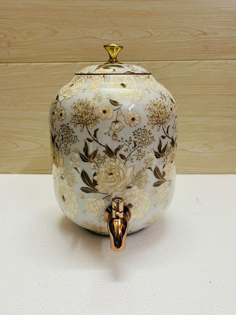 K&ouml;nnte beinhalten: Ein wei&szlig;er Getr&auml;nkespender aus Keramik mit einem floralen Muster in Gold und Braun. Er hat einen goldenen Deckel und einen kupferfarbenen Zapfhahn. Geeignet zum Servieren von Getr&auml;nken.