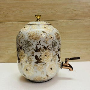 K&ouml;nnte beinhalten: Ein wei&szlig;er Getr&auml;nkespender aus Keramik mit einem floralen Muster in Gold und Braun. Der Spender hat einen Deckel mit einem goldenen Knopf und einen Wasserhahn aus Bronze. Das florale Design umfasst Rosen und andere Blumen.