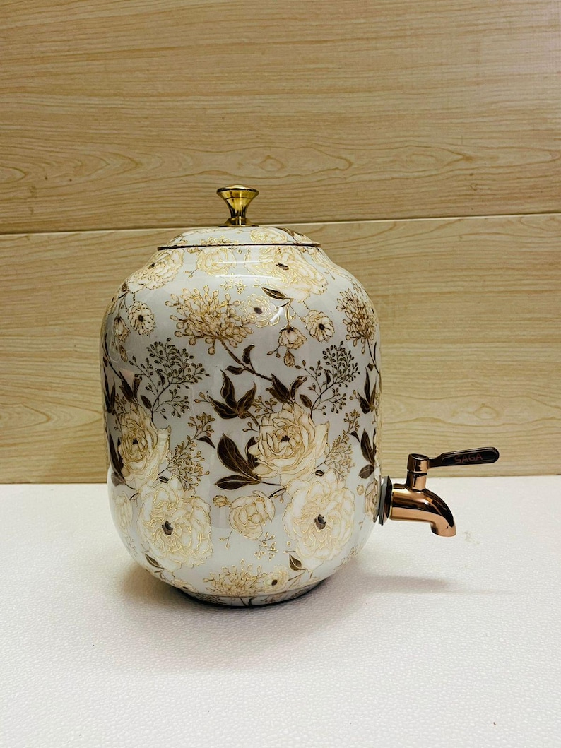 K&ouml;nnte beinhalten: Ein wei&szlig;er Getr&auml;nkespender aus Keramik mit einem floralen Muster in Gold und Braun. Der Spender hat einen Deckel mit einem goldenen Knopf und einen Wasserhahn aus Bronze. Das florale Design umfasst Rosen und andere Blumen.