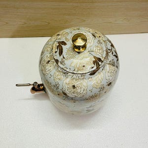 K&ouml;nnte beinhalten: Eine runde Teekanne mit floralem Muster. Die Teekanne hat einen hellblauen Hintergrund mit goldenen und wei&szlig;en Blumenmustern. Sie hat einen Deckel mit einem goldenen Knopf und einen Kupferausguss. Durchmesser ca. 20 cm.