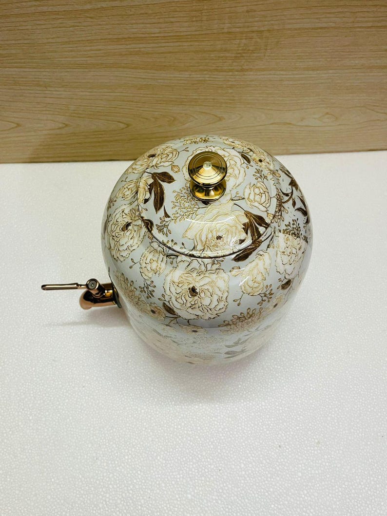 K&ouml;nnte beinhalten: Eine runde Teekanne mit floralem Muster. Die Teekanne hat einen hellblauen Hintergrund mit goldenen und wei&szlig;en Blumenmustern. Sie hat einen Deckel mit einem goldenen Knopf und einen Kupferausguss. Durchmesser ca. 20 cm.