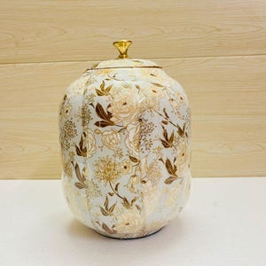 K&ouml;nnte beinhalten: Dekorative Keramikdose mit Deckel, mit hellblauem Hintergrund und einem floralen Muster in Gold und Braun. Die Dose hat eine abgerundete Form und einen goldfarbenen Knopf oben.