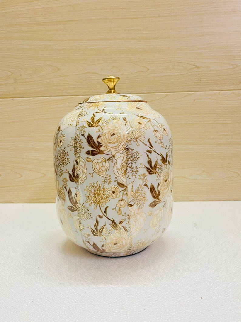 K&ouml;nnte beinhalten: Dekorative Keramikdose mit Deckel, mit hellblauem Hintergrund und einem floralen Muster in Gold und Braun. Die Dose hat eine abgerundete Form und einen goldfarbenen Knopf oben.