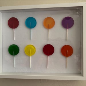 May include: A white rectangular shadow box frame containing eight round, translucent lollipops arranged in two rows of four. The lollipops feature distinct colors including red, blue, orange, purple, green, yellow, dark red, and reddish-orange.